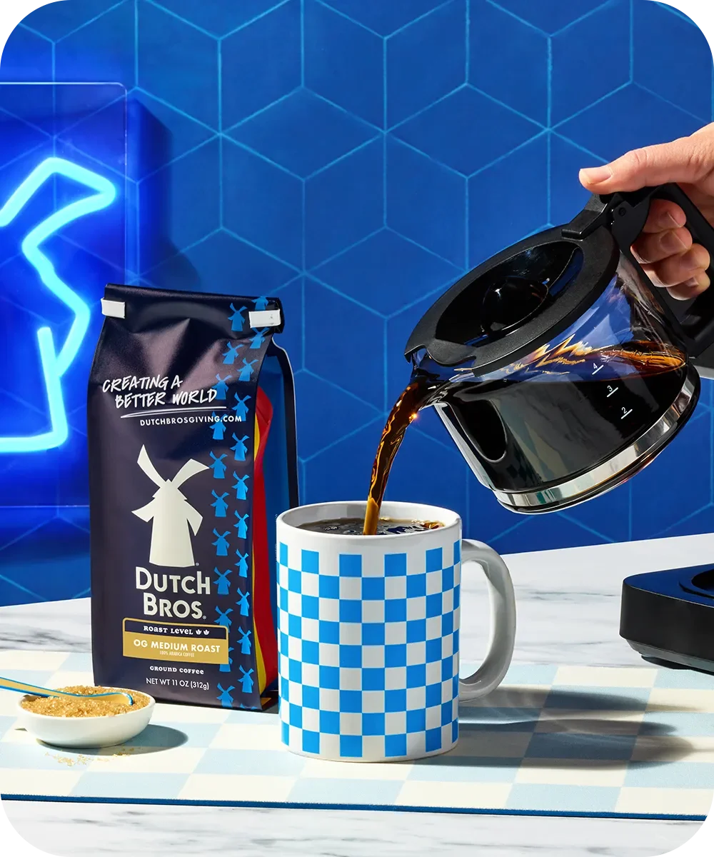 Hand pouring coffee from carafe into blue checkered mug, with Dutch Bros coffee bag against blue geometric background.