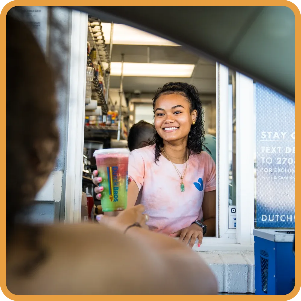Smiling server in pink shirt at counter window handing a colorful smoothie drink to a customer at Dutch Bros.