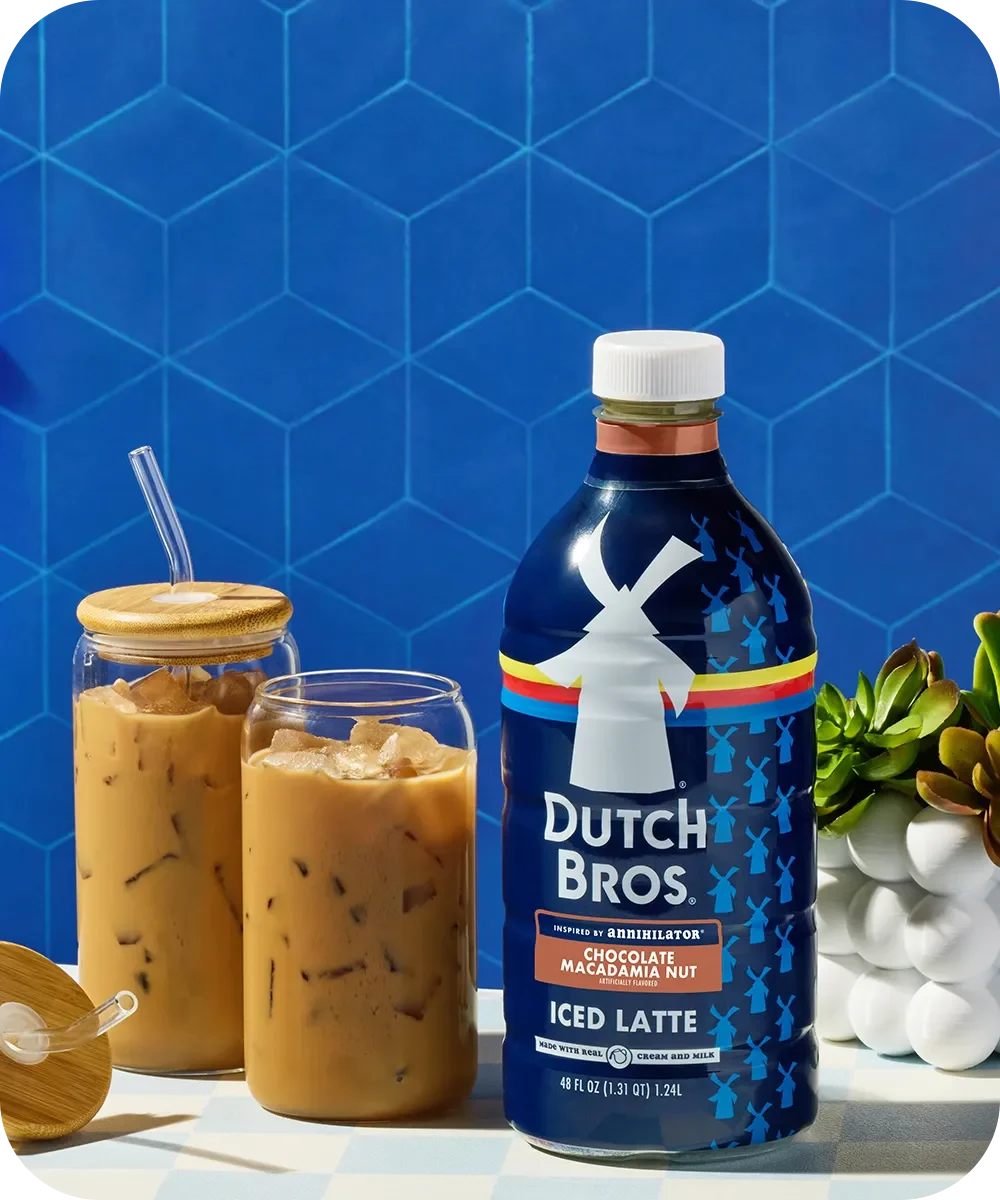 Dutch Bros Chocolate Macadamia Nut Iced Latte bottle with two glasses of iced coffee against a blue geometric background.
