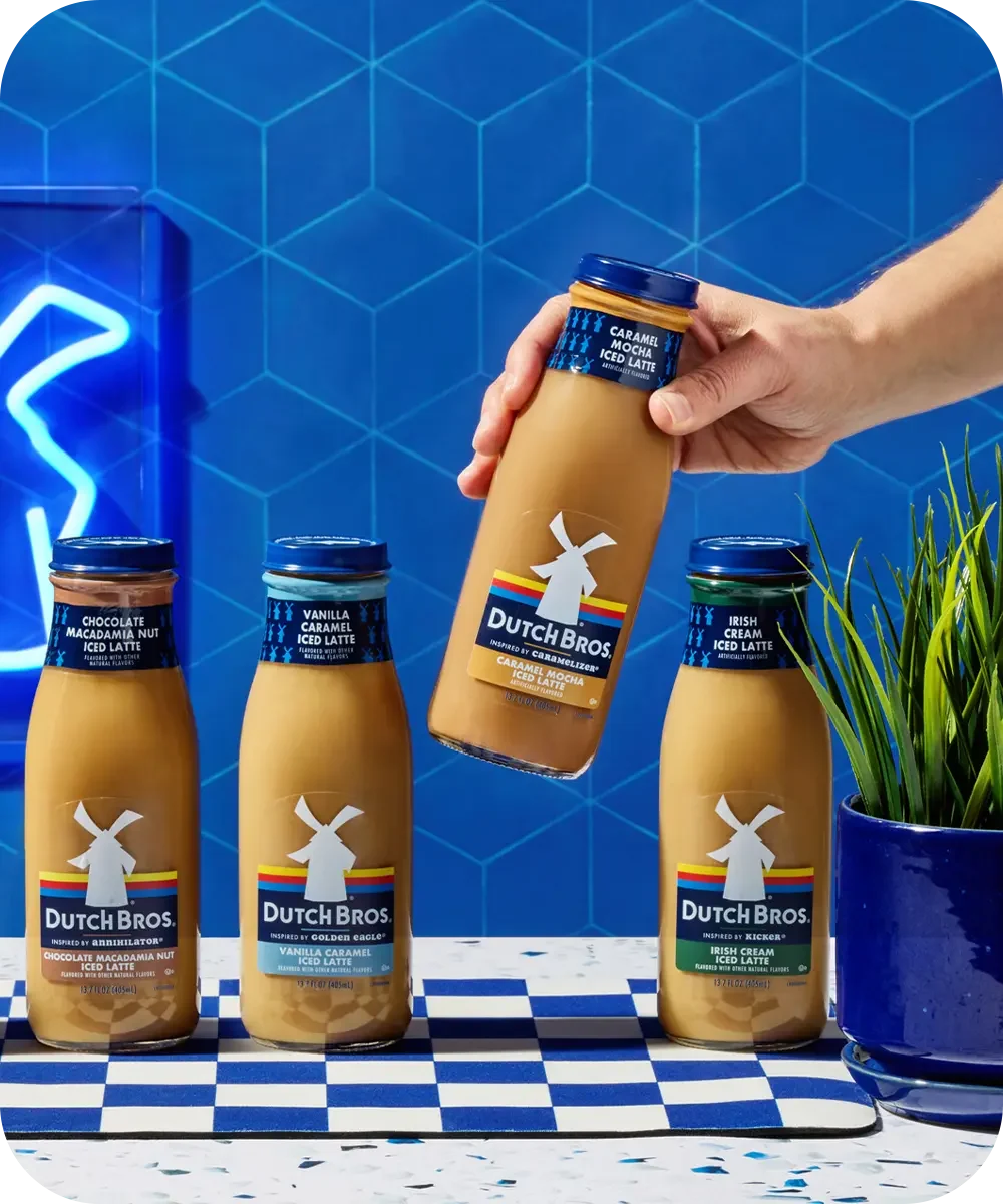 Hand holding Dutch Bros caramel iced latte bottle above three other flavored coffee bottles against blue tile background.