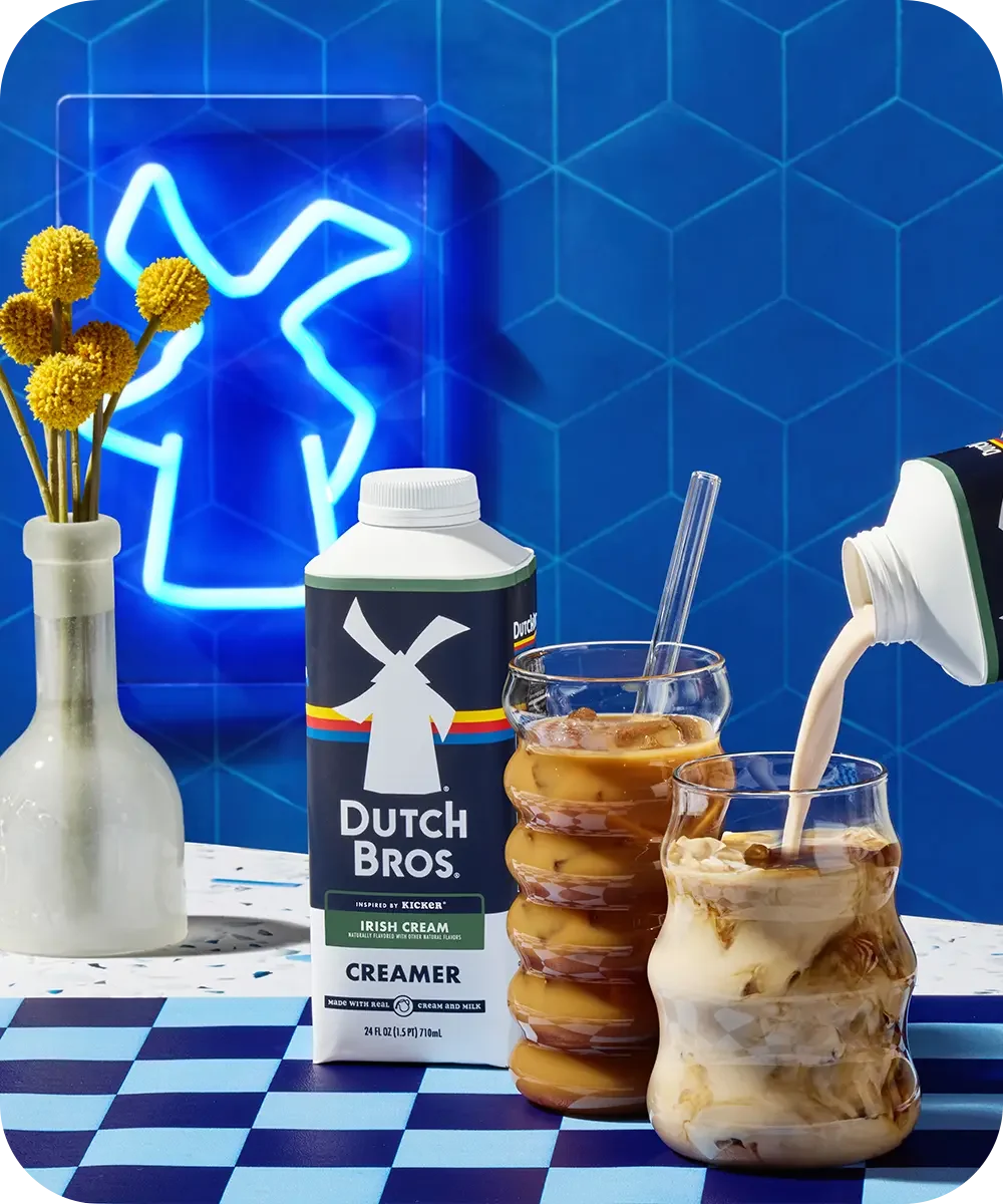 Dutch Bros Irish Cream creamer being poured into iced coffee drinks against blue tiled wall with neon light sign.