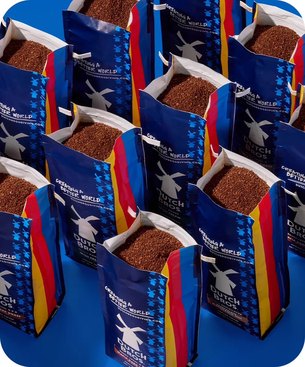 Multiple blue coffee bags with windmill logo and colorful stripes containing ground coffee, labeled "Creating A Better World"