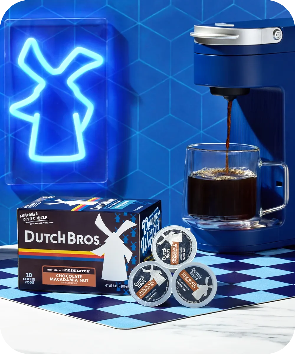 Blue coffee machine brewing Dutch Bros Chocolate Macadamia Nut coffee with product box and pods on checkered surface.