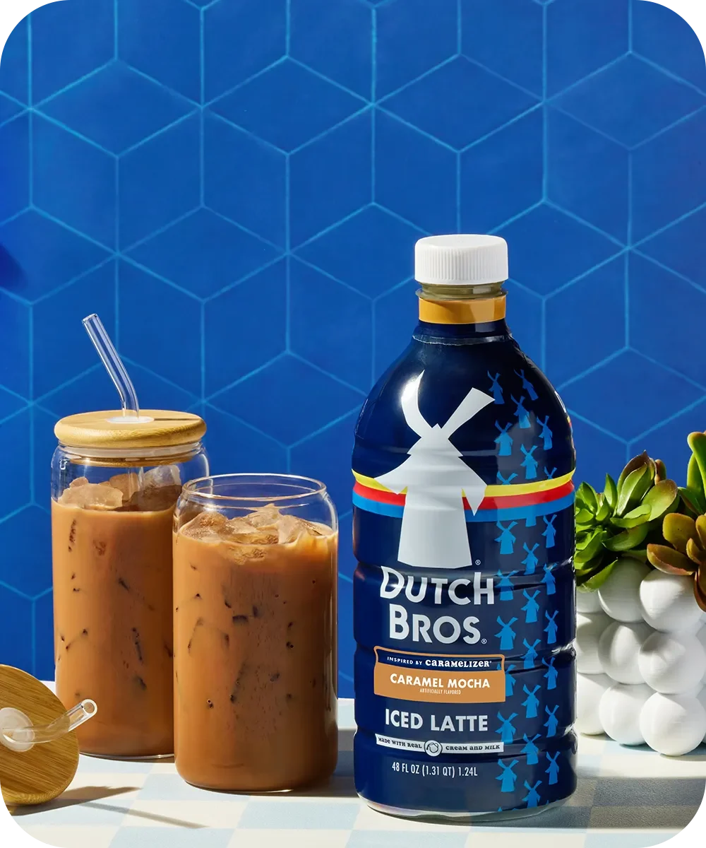 Dutch Bros Caramel Mocha bottle next to two glasses of iced coffee with a blue hexagon-patterned background.