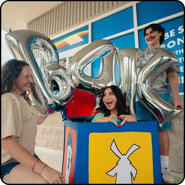 Three young people celebrating outdoors with large silver letter balloons spelling "B4K" near a colorful blue building.