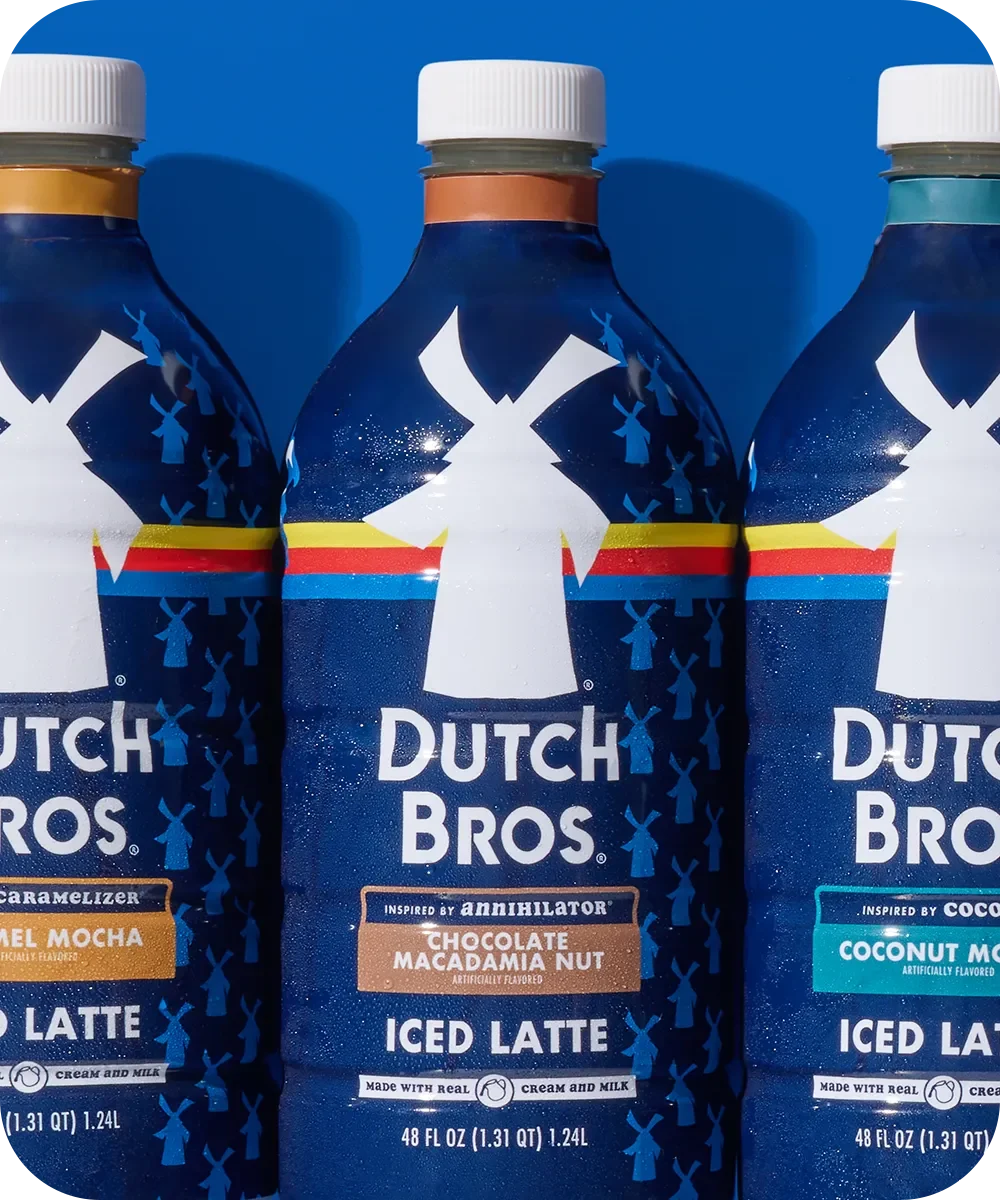 Three Dutch Bros bottled iced lattes with different flavors in dark blue bottles against a blue background.