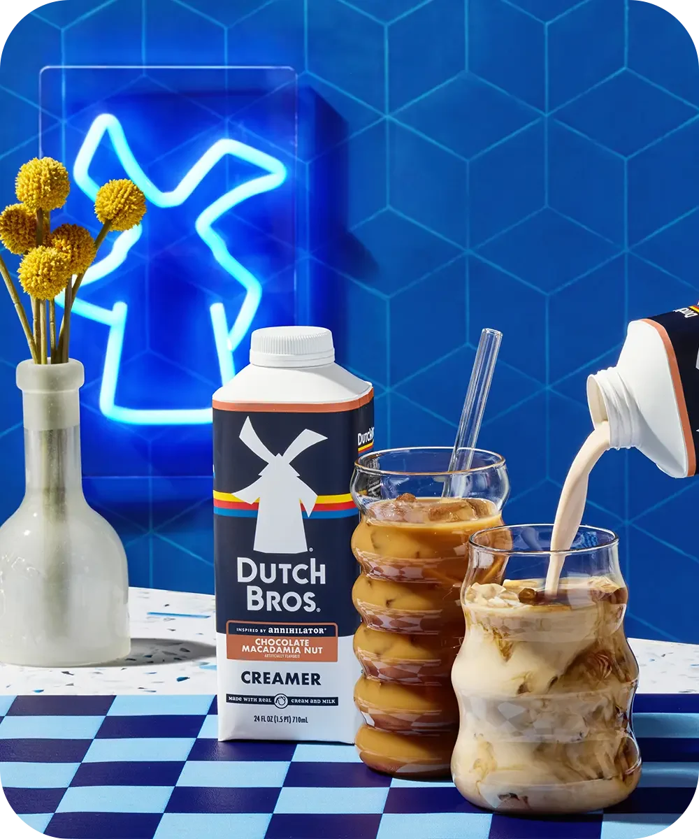 Dutch Bros chocolate macadamia creamer with iced coffees against blue tiled wall with neon sign and yellow flowers.
